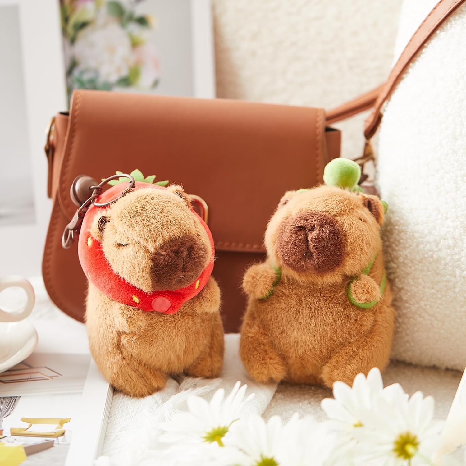 Two brown capybara plush keychains, one wearing a strawberry hood and one with a turtle backpack