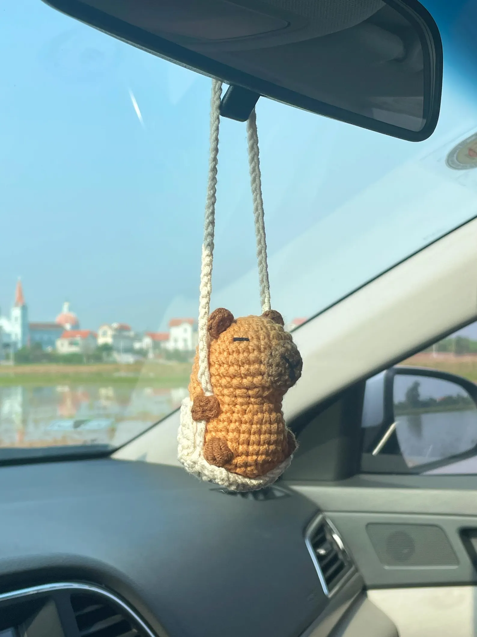 Crochet capybara hanging from a car rearview mirror, handmade in brown cotton yarn, sitting in a small yarn swing