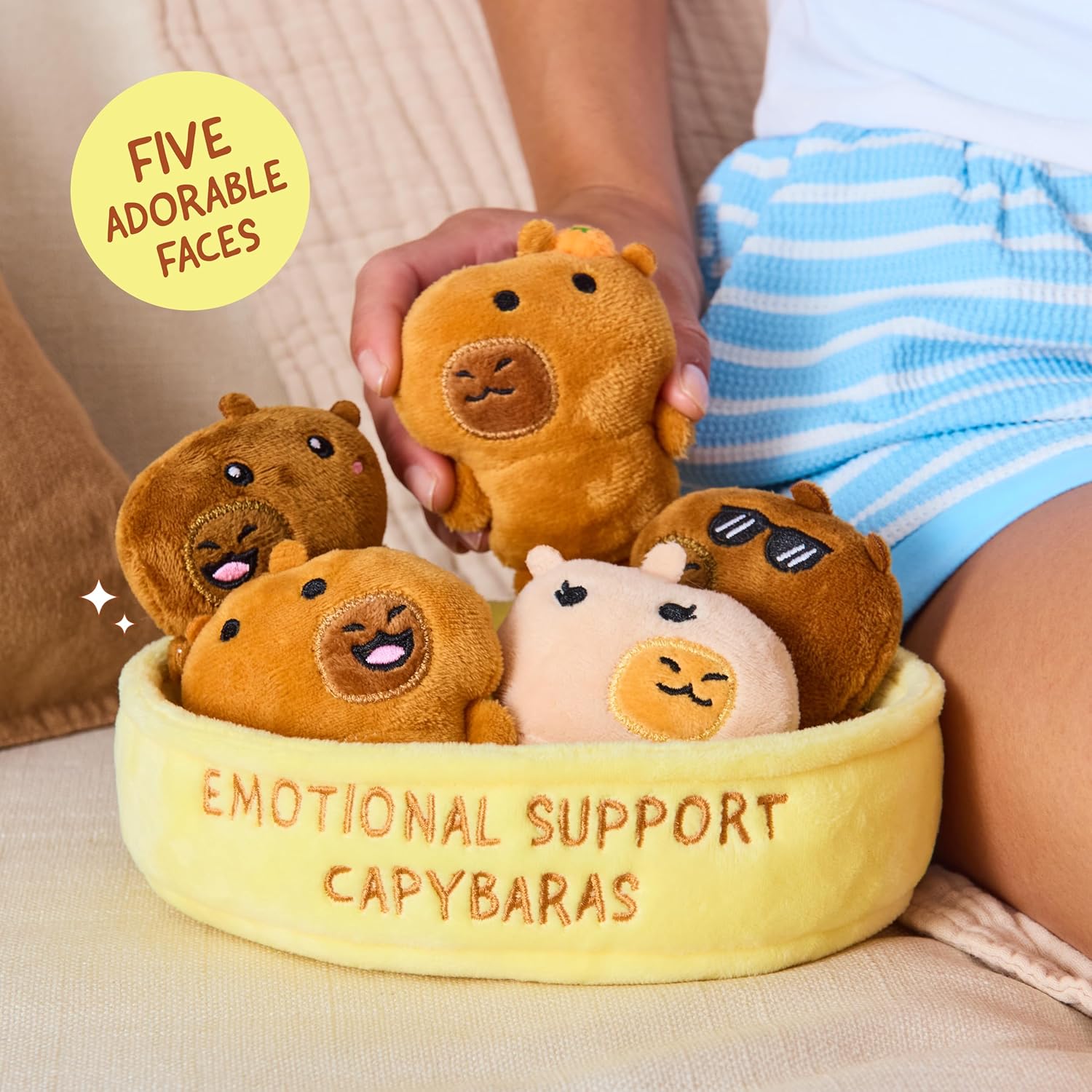 Five small capybara plush toys with different expressions sitting in a yellow felt basket labeled Emotional Support Capybaras, held by a person